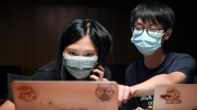 Junge Frau und junger Mann mit Schutzmasken sitzen vor 2 Monitoren