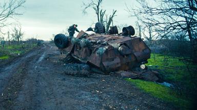 Das Bild zeigt eine trostlose Landschaft, die stark von den Folgen eines Krieges geprägt ist. Im Vordergrund liegt ein umgestürzter Panzer, dessen rostige Oberfläche und beschädigte Teile deutlich sichtbar sind. Auf dem Panzer steht ein Mann, der an dem Fahrzeug arbeitet. Seine Körperhaltung deutet auf eine gewissenhafte Beschäftigung hin. Im Hintergrund sind weitere, kahle Bäume sichtbar, die kaum noch lebendes Geäst tragen. Der Himmel ist bewölkt und erscheint düster, was die traurige Atmosphäre der Szene verstärkt. Die Erde ist matschig und wirkt unordentlich, mit verstreuten Überresten, die auf die Zerstörung in der Umgebung hinweisen. Der Weg, der sich durch die Landschaft schlängelt, ist ebenfalls von Schlamm und Trümmern gezeichnet. Alles in diesem Bild vermittelt ein starkes Gefühl von Verwüstung und den Auswirkungen des Krieges auf das tägliche Leben der Menschen in der Region.