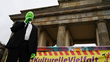 Mann mit grüner Strumpfmaske vorm Gesicht steht vorm Brandenburger Tor.