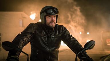 "Dengler - Am zwölften Tag": Georg Dengler (Ronald Zehrfeld) sitzt in Motorradkluft auf seinem Motorrad, im Hintergrund sieht man eine brennende Scheune.