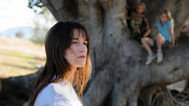 Eine Frau blickt an der Kamera vorbei. Im Hintergrund klettern Kinder auf einem Baum.