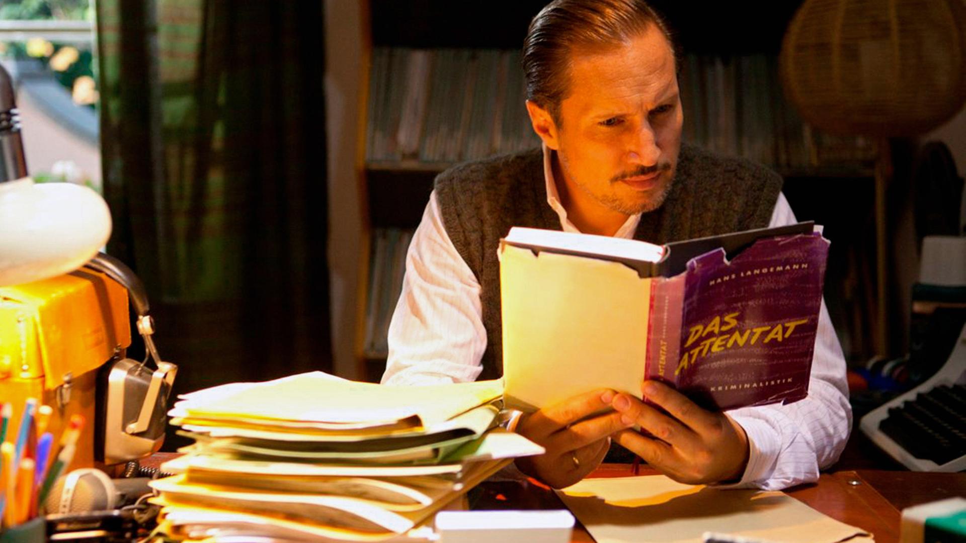 Ulrich Chaussy (Benno Fürmann) sitzt am Schreibtisch und liest das Buch "Das Attentat".