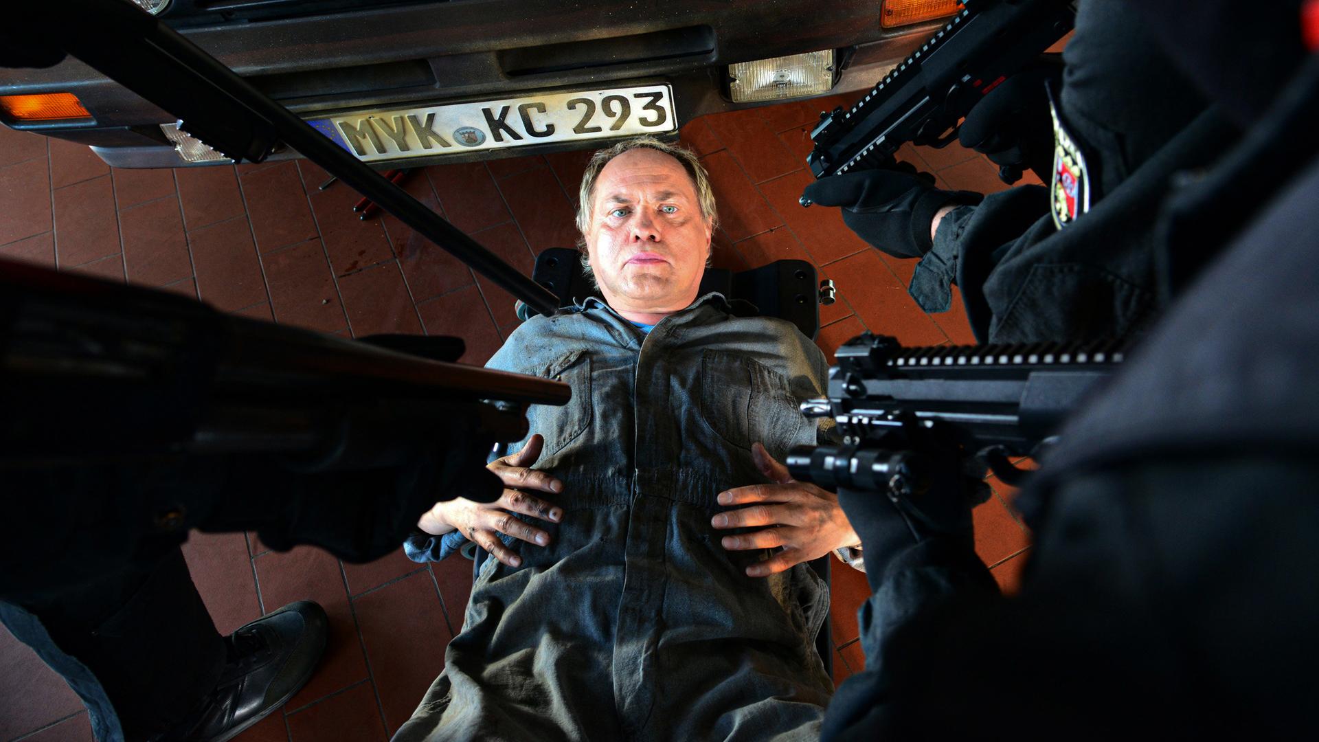 Ein Mechaniker liegt auf dem Boden, hinter ihm ein Auto, über ihm werden Pistolen auf ihn gerichtet.