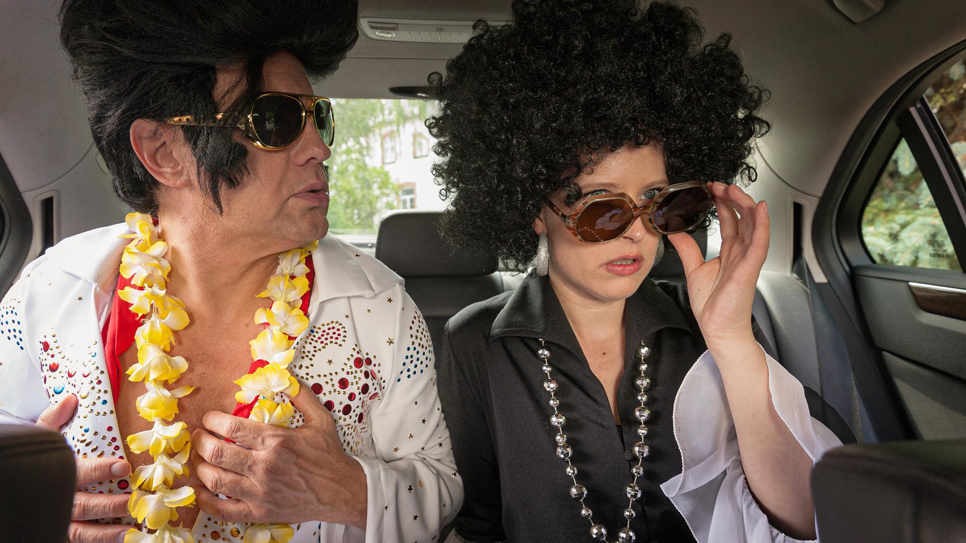 Zwei verkleidete Menschen mit Perücken sitzen in einem Auto