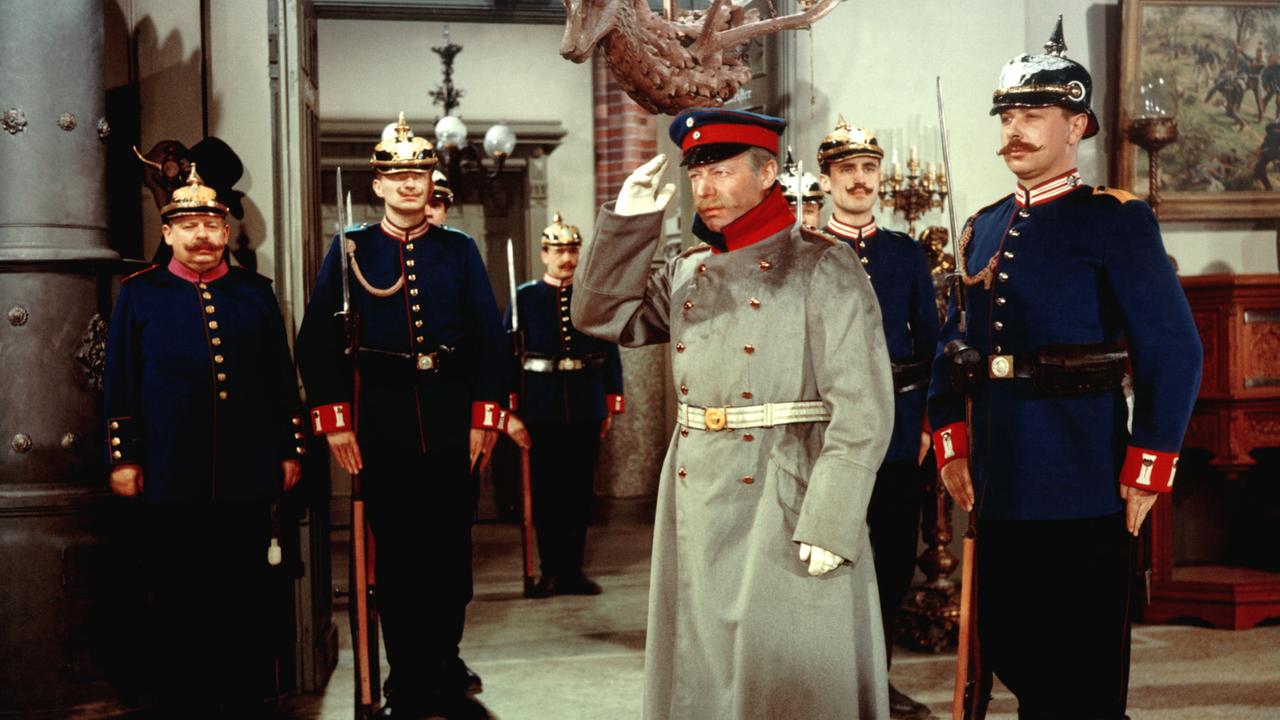 Ein Mann in langem Offiziersmantel steht militärisch grüßend in einem repräsentativen Raum, hinter sich ein sechs Mann starker Trupp von Soldaten in blauem Uniformrock und mit Pickelhaube.