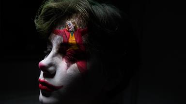 Nahaufnahme eines als Clown geschminkten Jungen. Sein Gesicht ist zur Hälfte im Dunkeln, auf seiner Stirn ist klein eine Szene des tanzenden Jokers eingeblendet.