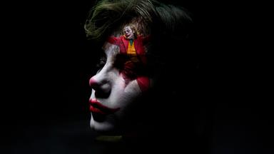 Nahaufnahme eines als Clown geschminkten Jungen. Sein Gesicht ist zur Hälfte im Dunkeln, auf seiner Stirn ist klein eine Szene des tanzenden Jokers eingeblendet.