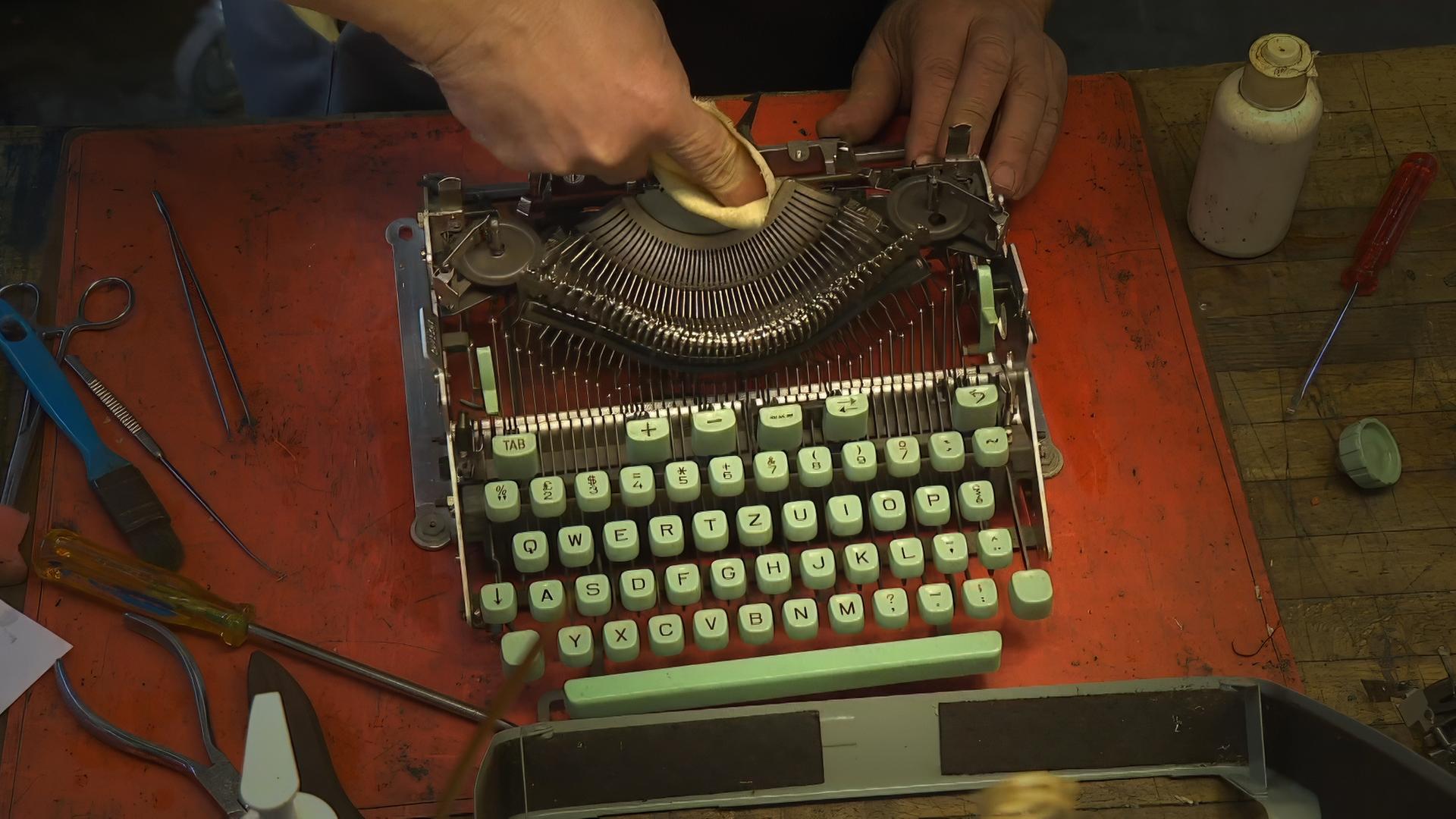 Nahaufnahme einer mechanischen Schreibmaschine mit mintgrünen Tasten, die mit einem Tuch im Innenbereich gereinigt wird; umgeben von verschiedenen Werkzeugen auf einer Werkbank.