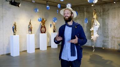 Ein blau gekleideter Mann mit Hut steht gestikulierend in einer Skulpturenausstellung