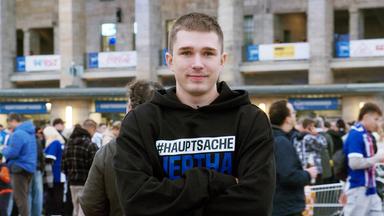 Lächelnder junger Mann mit verschränkten Armen vor dem Berliner Olympia-Stadion.