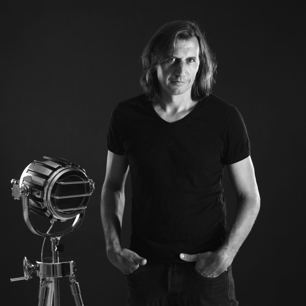 Schwarzweißfoto: Klaus Stern, bekleidet mit schwarzer Jeans und schwarzem T-Shirt, hat die Hände halb in die Hosentaschen geschoben und steht vor einem schwarzen Hintergrund. Er schaut mit offenem Blick in die Kamera, neben ihm ein kleiner, metallisch glänzender Studioscheinwerfer als Requisite.