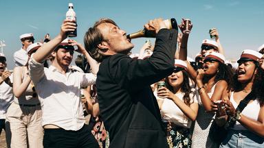 Ein Mann trinkt aus einer Flasche. Um ihn herum jubelnde dänische Schulabsolventen.