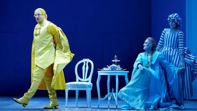 Ein Schauspieler auf einer blauen Bühne in gelber Kleidung läuft an zwei Schauspielerinnen vorbei.