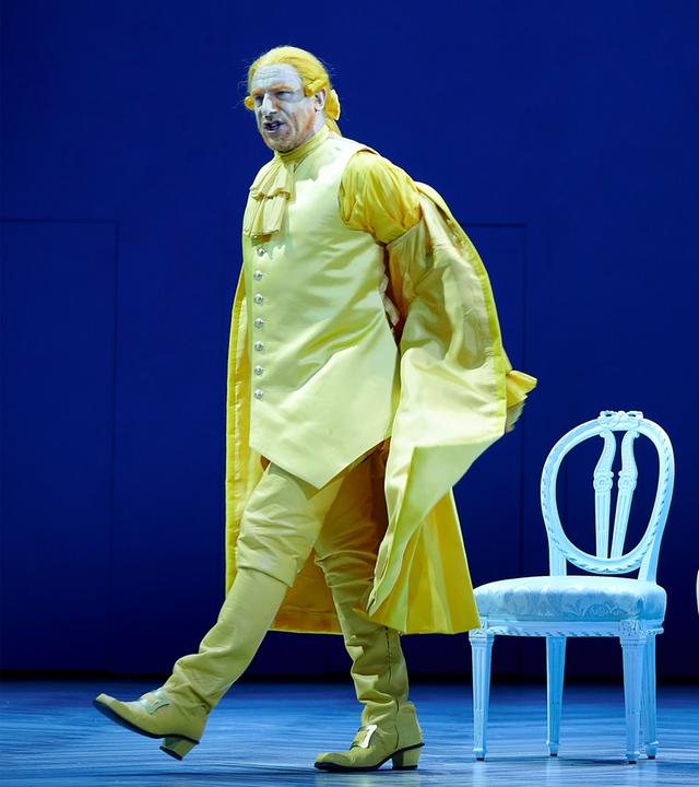 Ein Schauspieler auf einer blauen Bühne in gelber Kleidung läuft an zwei Schauspielerinnen vorbei.