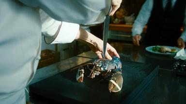 Ein Koch hat ein spitzes Küchenmesser hinter dem Kopf eines Hummers angesetzt, der vor ihm auf einem Tisch liegt.