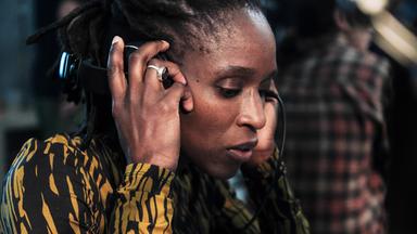 Die Astrophysikerin Samantha Crowe (Sharon Duncan-Brewster) trägt Kopfhörer, die sie mit beiden Händen auf ihre Ohren drückt. Sie schaut konzentriert zu Boden.