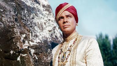 Der Maharadscha von Eschnapur (Walther Reyer) trägt eine herrschaftliche weiße Tracht mit einer prunkvollen Halskette, einer roten Schärpe sowie einem roten Turban und lehnt wie in Pose stehend an einem Felsen. Sein skeptisch-versonnen wirkender Blick richtet sich nach schräg oben.