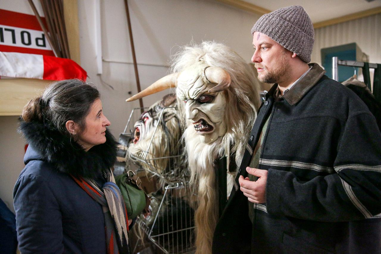 Ein Mann mit Mütze und eine Frau im Anorak sehen sich an. Zwischen ihnen ist eine Horror-Maske mit Hörnern und zottligem Haar zu sehen. 