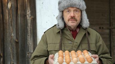 Ein Mann mit Fellmütze und Anorak vor einer Scheune hält einen Karton mit Eiern in der Hand.