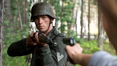 Walter Proska (Jannis Niewöhner) steht in Militäruniform und Gewehr im Wald und zielt auf eine Person, die eine Pistole auf ihn hält