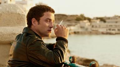 Ein Mann, der eine Zigarette hält, blickt nachdenklich auf eine Stadtansicht am Wasser in Marseille.