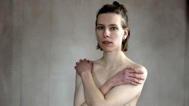 Nele steht mit vor der Brust verschränkten Armen vor einer Wand und schaut in die Kamera.