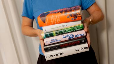 Eine Person hält einen Bücherstapel so vor sich, dass die Titel auf den Buchrücken zu lesen sind.