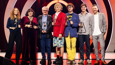 Es stehen sieben Personen auf einer Bühne. Der Hintergrund ist rot beleuchtet. Die Personen halten Trophäen in den Händen.