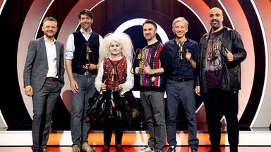 Es stehen sechs Personen auf einer Bühne. Der Hintergrund ist rot und weiß beleuchtet. Die Personen halten Trophäen in den Händen.