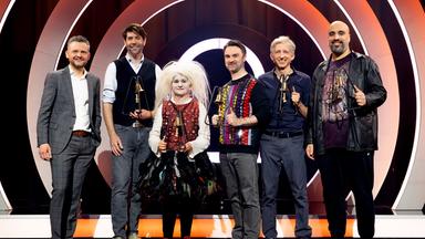 Es stehen sechs Personen auf einer Bühne. Der Hintergrund ist rot und weiß beleuchtet. Die Personen halten Trophäen in den Händen.