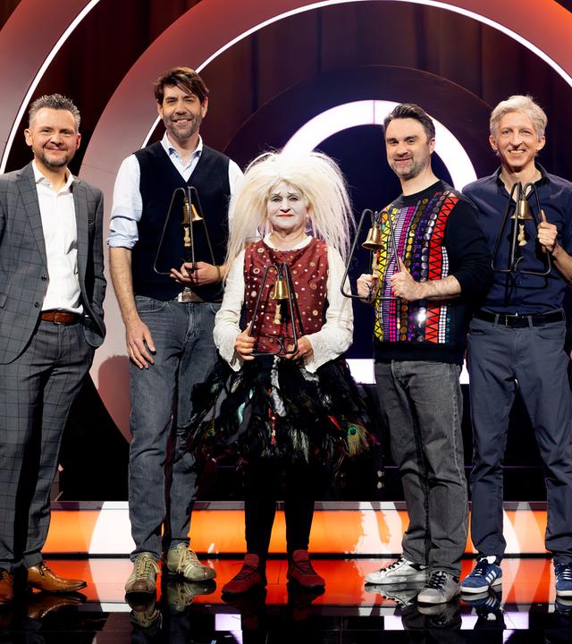 Es stehen sechs Personen auf einer Bühne. Der Hintergrund ist rot und weiß beleuchtet. Die Personen halten Trophäen in den Händen.