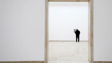 Blick durch eine große Türöffnung in einen großen weißen Ausstellungsraum, an dessen hinterer Wand ein großer QR-Code hängt, vor dem eine Person steht.