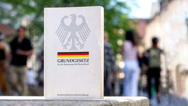 Das deutsche Grundgesetz steht in einer Einkaufsstraße, im Hintergrund sind verschwommen Menschen zu sehen.
