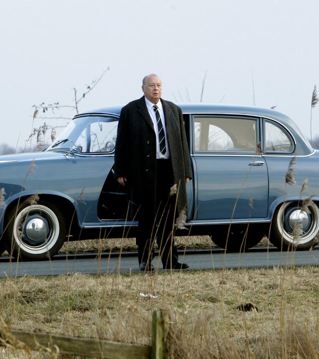 Mann steht vor seinem Oldtimer auf einer Landstraße.