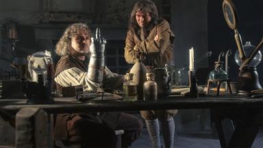 "Die glorreichen 10 - Die allerkrassesten Abstürze der Geschichte": Spielszene: Götz von Berlichingen sitzt an einer Werkbank, seinen rechten Arm mit der eisernen Hand auf die Werkbank gestützt. Von den Fingern dieser Hand ist nur der Mittelfinger ausgestreckt. Neben ihm steht ein Mann, der seine Arme vor der Brust verschränkt hat und nachdenklich auf die eiserne Hand schaut.