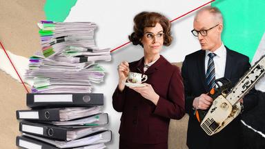 Maike Kühl und Max Uthoff stehen neben einem großen Stapel Aktenordner. Maike Kühl hält eine Tasse Tee in der Hand, Max Uthoff hält eine Motorsäge.