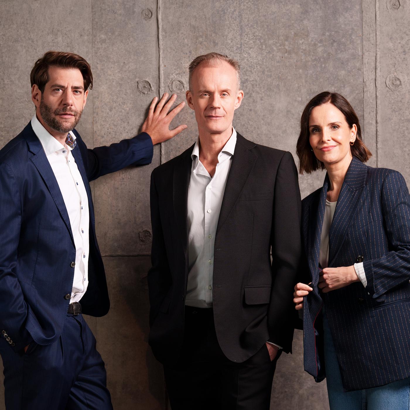 Drei Personen in Business-Kleidung stehen vor einer grauen Betonwand und blicken in die Kamera; links ein Mann im blauen Anzug mit einer Hand an der Wand, in der Mitte ein Mann im schwarzen Anzug, rechts eine Frau im dunkel gestreiften Blazer.