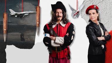 Max Uthoff und Maike Kühl stehen mit verschränkten Armen da und an Fäden hängen Bilder von Patronen, einem Kampfflugzeug und einer blutbespritzten Friedenstaube von der Decke. 