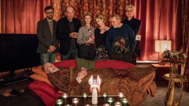 Menschen stehen um einen auf dem Sofa liegenden alten Mann versammelt.