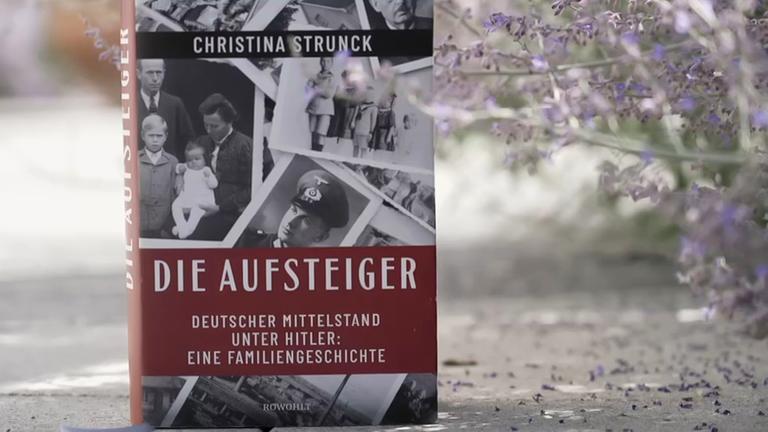 Draußen auf einer asphaltierten Straße steht vor lila Blütenzweigen ein Buch mit dem Titel "Die Aufsteiger" und alten Familienfotos auf dem Boden.