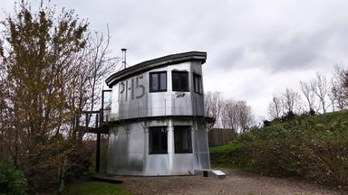 Ein silberner, zweistöckiger Wachturm mit großen Fenstern und der Nummer 'PH15' an der Seite steht an einem Weg im Grünen. Er ist umgeben von Bäumen und bewölktem Himmel