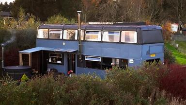 Ein blauer Doppeldeckerbus wurde in ein Haus umgebaut. Er steht im Grünen und hat mehrere Fenster, einem Schornstein und einen überdachten Eingang.