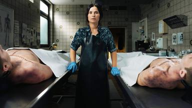 Eine Frau steht zwischen zwei aufgebarten Leichen.