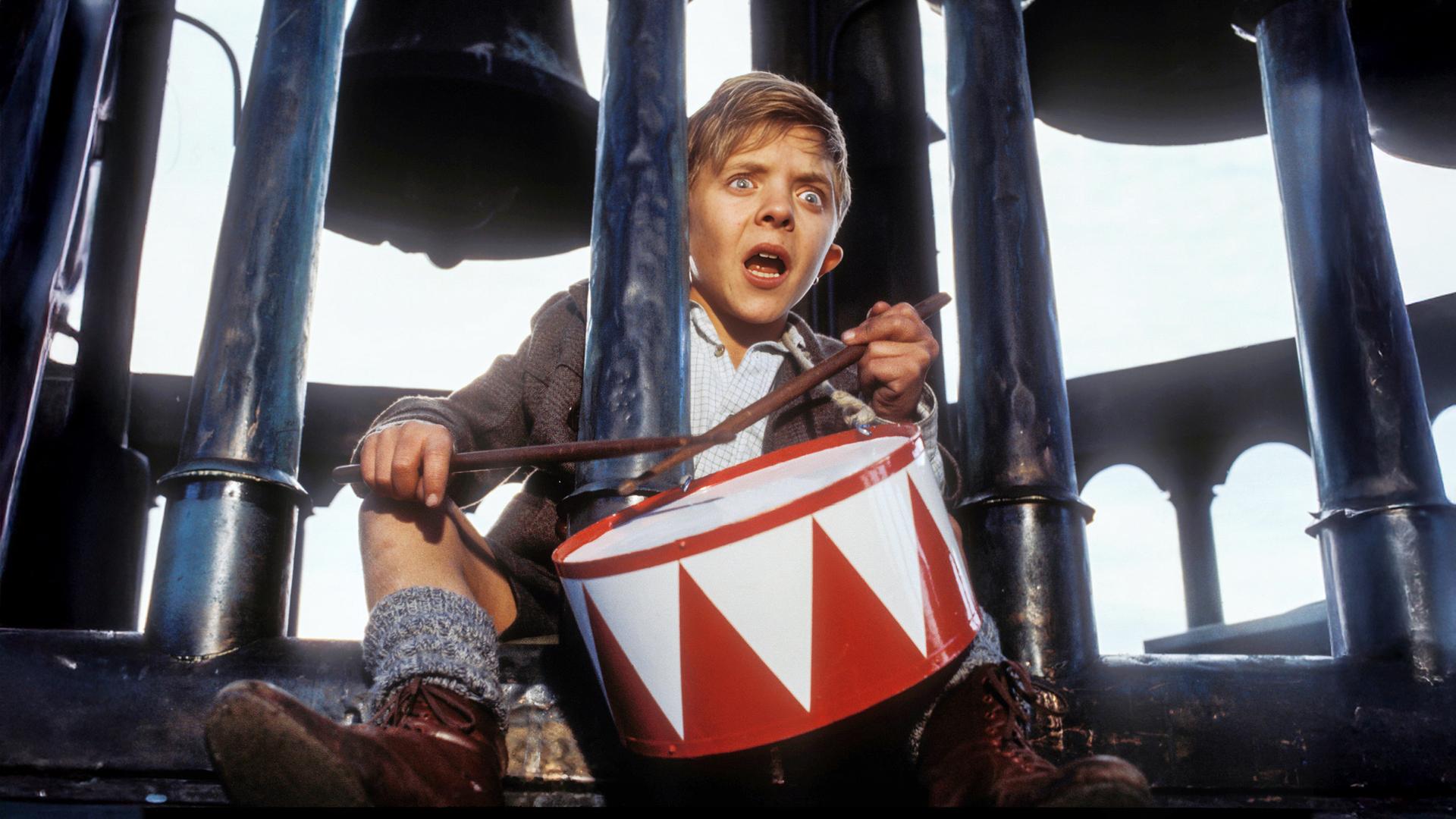 Ein Junge mit erschrockenem Blick sitzt auf einem Glockenturm und schlägt mit zwei Stöcken auf eine rot-weiße Spielzeugtrommel.