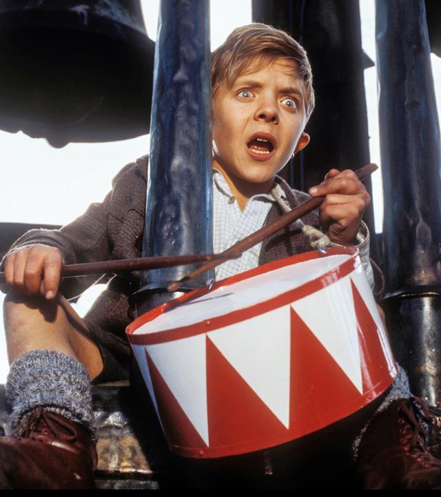 Ein Junge mit erschrockenem Blick sitzt auf einem Glockenturm und schlägt mit zwei Stöcken auf eine rot-weiße Spielzeugtrommel.