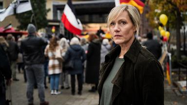 Claudia Voss (Anna Schudt) steht am Rande eines öffentlichen Platzes, auf dem sich mehrere Menschen, einige mit Fahnen, versammelt haben, und schaut nachdenklich um sich.