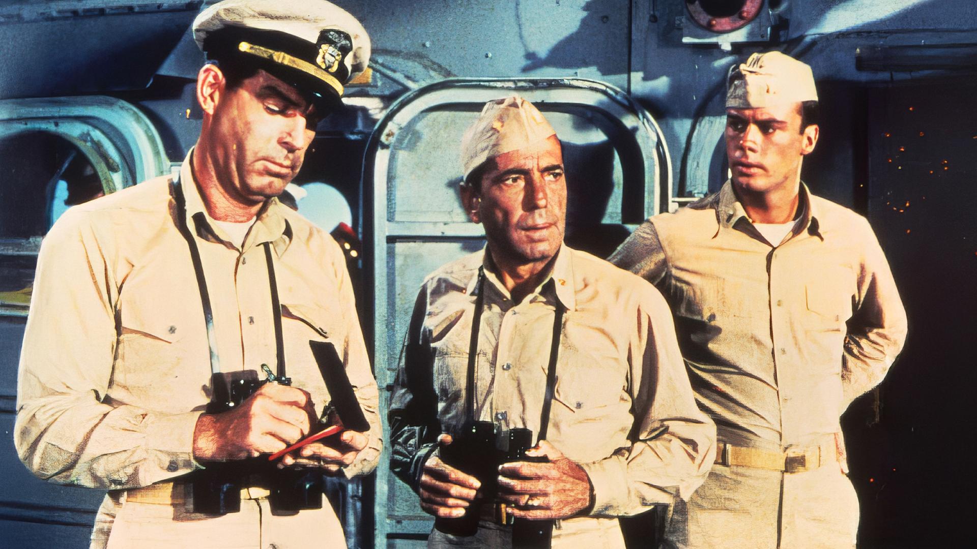 Vor den Aufbauten eines Schiffes steht ein Mann in Uniform  (Humphrey Bogart) mit einem umgehängten Fernglas. Rechts und links von ihm befinden sich zwei weitere Uniformierte, von denen der Mann links Eintragungen in eine Art Buch vornimmt, während der andere erstaunt blickt und die Hände in die Seiten stemmt.