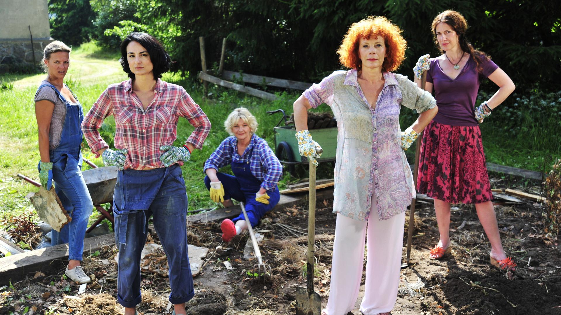 Fünf Frauen stehen in Arbeitskleidung und mit Gartengeräten in einem Gemüsebeet. Im Hintergrund sieht man eine Wiese, Bäume und den Teil einer Hausmauer.