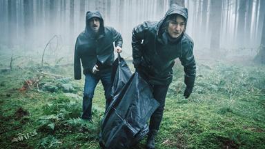 Ein Mann und eine Frau tragen einen Leichensack durch den Wald.