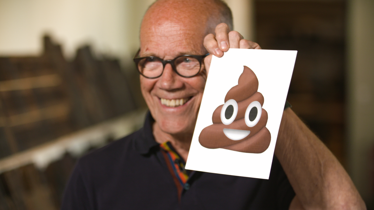 Mann mit Brille und schütterem Haar hält lachend ein Papier mit einem Kack-Emoji in die Luft.